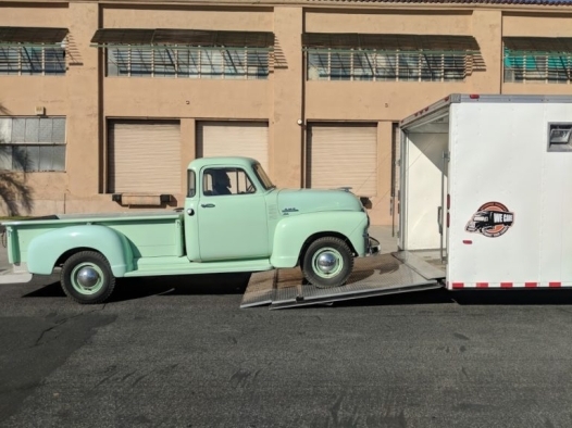 A light green GMC pickup truck is halfway loaded on the ramp into a white trailer with a WE CARE logo.