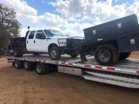 White pickup truck with flatbed and toolbox loaded on a trailer.