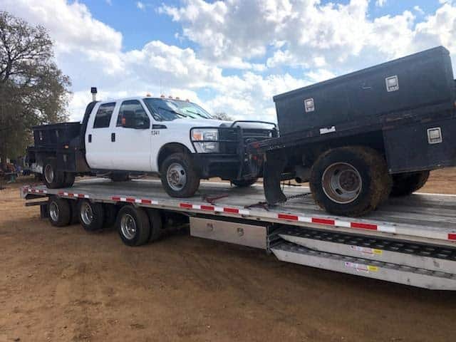 White pickup truck with flatbed and toolbox loaded on a trailer.