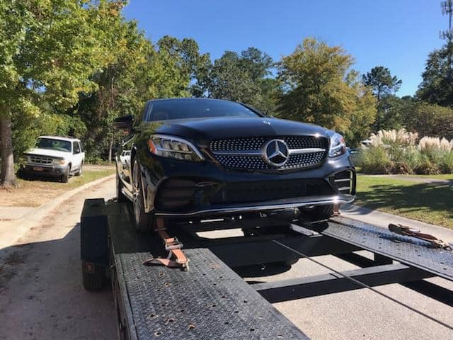 A black Mercedes-Benz car strapped onto a tow trailer, in front of a white pickup truck and lush green trees.