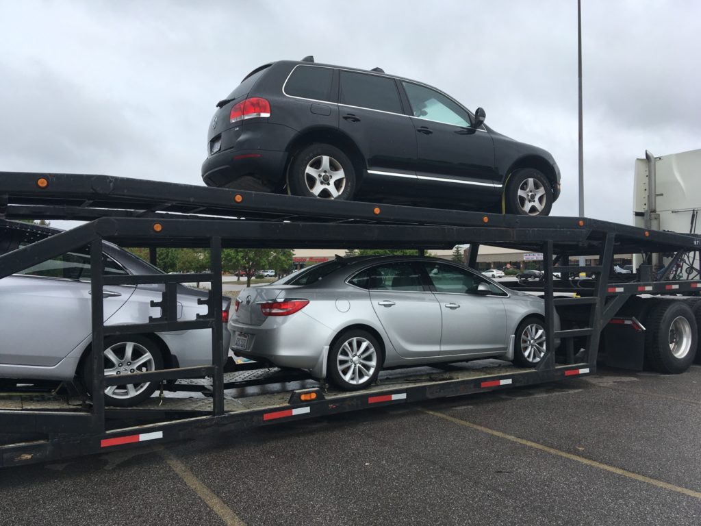 A car carrier truck is transporting three cars, including a black SUV on the top rack and two silver sedans on the bottom rack.