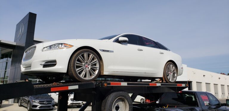 White Jaguar XJ on a black flatbed truck in front of a Mercedes-Benz dealership.