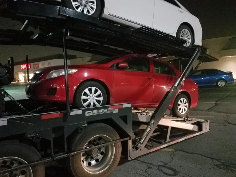 Red car and white car on a vehicle transport trailer in a parking lot.