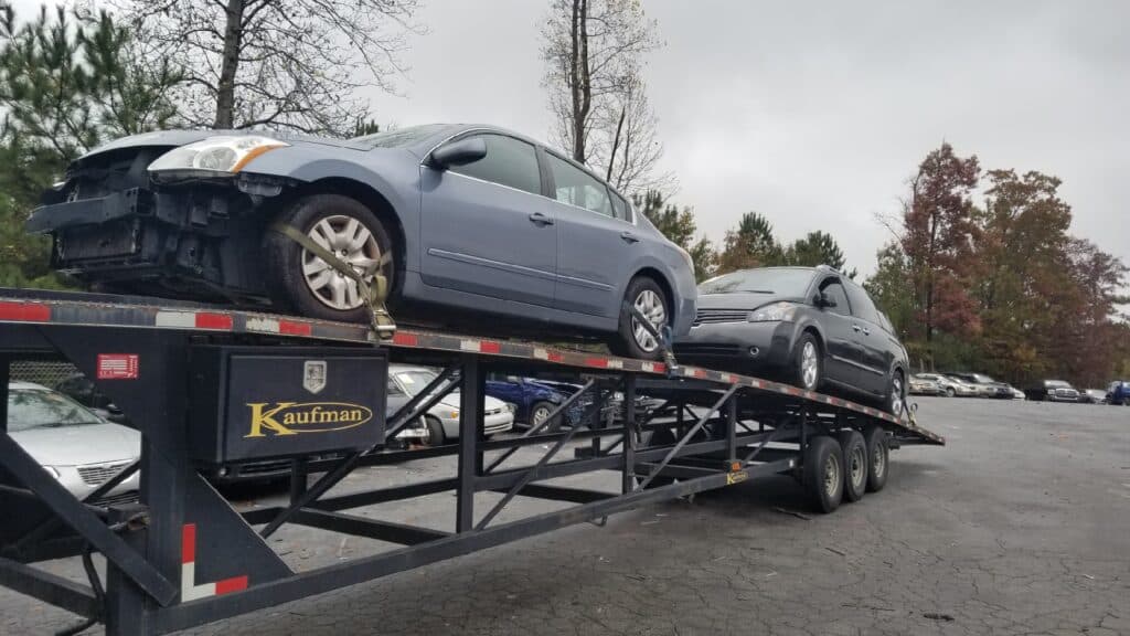 Two cars being hauled on a car carrier trailer on a cloudy day, with Kaufman branding on a black box on the side of the trailer.